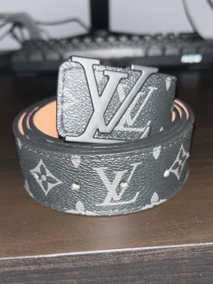 Svart L0uis Vuton monogram skärp - Snyggt svart skärp från L0uis Vuton med klassiskt monogrammönster och stor L&V-logga i metall som spänne. Skärpet har en lyxig känsla och passar perfekt till jeans eller kostymbyxor. Materialet är skinn med detaljer i silver.