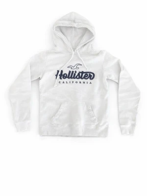 Vit hoodie från Hollister - Snygg vit hoodie från Hollister med broderad logga i mörkblått på bröstet. Klassisk känguruficka framtill och justerbar huva med dragsko. Perfekt för en avslappnad och trendig stil.