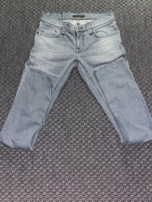 Gråa jeans från Nudie Jeans - Säljer ett par gråa jeans från Nudie Jeans med klassisk femficksmodell och snygga vågdetaljer på bakfickorna. Jeansen har normal midja, raka ben och är tillverkade i mjuk denim. Perfekta för en avslappnad och trendig stil.