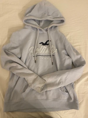 Ljusblå hoodie från Hollister - Snygg ljusblå hoodie från Hollister med broderad logga och text på bröstet. Klassisk känguruficka framtill, justerbar huva med dragsko och ribbade muddar. Perfekt för en avslappnad och trendig look.