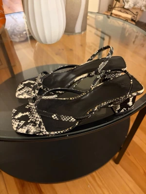 Svarta klackar med ormmönster - Säljer ett par trendiga klackar med svart och vitt ormmönster, fyrkantig tå och låg blockklack. Remmarna är tunna och eleganta, och sulan är svart. Perfekta för dig som vill sticka ut med en cool look. Materialet känns som konstläder.