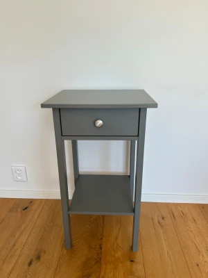 Grått sängbord (Mio) med låda och hylla - Snyggt grått sängbord i trä med en låda och en öppen hylla längst ner. Bordet har raka ben och en rund metallknopp på lådan. Perfekt för att förvara småprylar och böcker bredvid sängen.