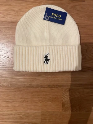 Vit ribbstickad mössa med Polo Ralph Lauren logga  - Snygg vit ribbstickad mössa med Polo Ralph Lauren logga broderade framtill. Tillverkad i mjukt material som håller dig varm och stilsäker under kalla dagar. Perfekt accessoar för dig som vill ha en clean och tidlös look.