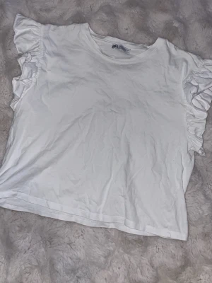 Vit volangtopp från Zara - Säljer en söt vit topp från Zara med volangdetaljer på ärmarna. Toppen har rund halsringning och är gjord i mjuk bomull, perfekt för en enkel men stylish look. Passformen är normal och toppen är kortare i modellen.