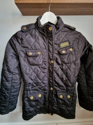 Svart quiltad jacka från Barbour - Cool svart quiltad jacka från Barbour med guldiga knappar och fyra fickor framtill. Jackan har hög krage, axelklaffar och ett snyggt Barbour-märke på bröstet. Perfekt för dig som vill ha en klassisk och tidlös look. Materialet är mjukt och lätt vadderat. Jackan är använd och små slitningar som en höndig person lätt fixar. En del av översta knappen saknas, ses på bild 