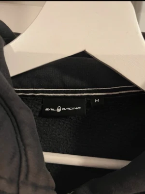 Svart hoodie från Sail Racing - Svart hoodie från Sail Racing med dragkedja framtill och två stora fickor. Klassisk huva och ribbade muddar vid ärmslut och nederkant. Diskret logga på bröstet. Perfekt för en avslappnad och sportig stil. Dock kan man se att tröjan är lite solblekt på ärmarna o nedtill tröjan 