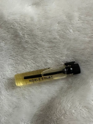 Noble Eclat parfymprov essnce - Litet parfymprov från Noble Eclat i genomskinlig glasflaska med svart skruvkork. Flaskan är cylinderformad och innehåller en ljusgul vätska. Perfekt att ha med i väskan eller testa en ny doft.