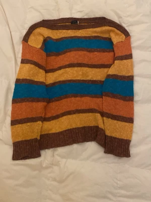 Randig stickad tröja från KappAhl - Färgglad stickad tröja från KappAhl med breda ränder i brunt, gult, orange och blått. Tröjan har rund halsringning, långa ärmar och ribbstickade muddar. Perfekt för dig som gillar retrovibbar och mjuka material.