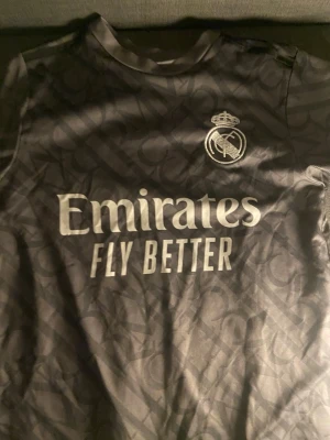 Real Madrid Bellingham matchtröja - Snygg svart Real Madrid matchtröja med Bellingham och nummer 5 på ryggen. Tröjan har klubbmärke och sponsortryck på bröstet samt diskret mönster i tyget. Perfekt för fotbollsfans som vill visa sitt stöd för laget.