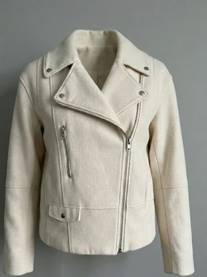 Krämvit kappa H&M XS - Snygg krämvit kappa i klassisk bikerstil med snedställd dragkedja, bred krage och silverfärgade detaljer. Jackan har en bröstficka med dragkedja. Perfekt för dig som vill ha en stilren och trendig look. Använd fåtal gånger, är i mycket bra skick. Storlek XS men passar även S, stor i storleken.