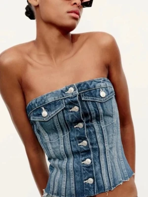 Blå denim tubtopp med knappar - Cool tubtopp i blå denim med knappar framtill och två dekorativa bröstfickor. Toppen har en bandeau-form och rå fransad nederkant som ger en edgy look. Perfekt för dig som vill sticka ut med en unik och trendig topp.