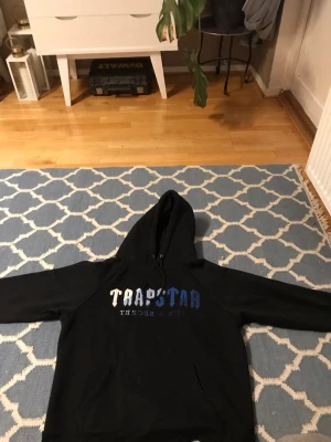Svart Trapstar hoodie  - Säljer en svart hoodie från Trapstar med blått och vitt tryck på bröstet där det står 'Trapstar' och 'It's a secret'. Klassisk huva med dragsko och känguruficka framtill. Perfekt för en avslappnad streetwear-look.
