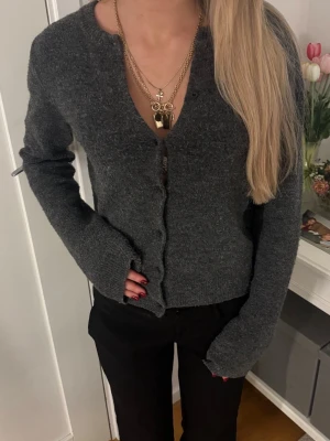 Mörkgrå stickad kofta med v-ringning - Mysig mörkgrå kofta med v-ringning och knäppning framtill. Långärmad modell i mjukt stickat material, perfekt att slänga på sig över en t-shirt eller linne. Enkel och stilren design som passar till det mesta.