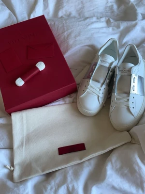 Valentino vita sneakers med silverdetalj - Snygga vita sneakers från Valentino med bred silverfärgad rem över mitten och diskret logga på plösen. Skorna har en klassisk låg siluett och är tillverkade i slätt läder. Perfekta för dig som vill ha en stilren men ändå lyxig look.