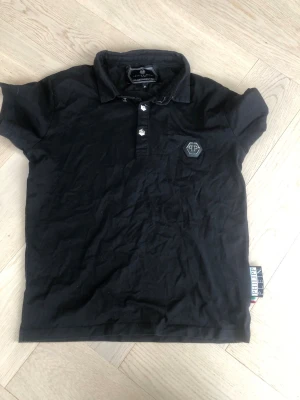 Svart polotopp från Philipp Plein - Cool svart polotopp från Philipp Plein med korta ärmar och klassisk krage. Toppens bröst pryds av ett metallic PP-emblem och den har knappar framtill. Perfekt för dig som gillar exklusiva märken och stilren design.