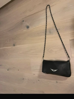 Svart axelväska från Zadig & Voltaire - Snygg svart axelväska i läder från Zadig & Voltaire med silverfärgad kedjerem och ikonisk vinge i metall framtill. Väskan har en rektangulär form och dragkedja nertill. Perfekt för dig som gillar en edgy och stilren look. Det korta bandet saknas tyvärr därför säljer jag den billigt