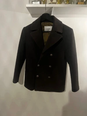  skepparkavaj från Dondup - Stilren mörkbrun skepparkavaj från Dondup med dubbelknäppning och bred krage. Klassisk rak passform och fyra dekorativa metallknappar framtill. Tillverkad i ett tjockare tyg som ger en snygg siluett och passar perfekt till höstens outfits.