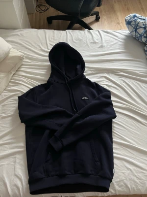 Mörkblå hoodie från Les Deux - Säljer en mörkblå hoodie från Les Deux med klassisk huva och dragsko. Tröjan har en liten broderad logga på bröstet och en stor magficka framtill. Tillverkad i mjukt bomullsmaterial som är skönt att ha på sig.