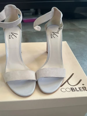 Beige mocka-pumps från NLY Shoes - Stilrena pumps i beige mocka från NLY Shoes. Skorna har öppen tå, bred rem över foten och justerbart ankelband med silverfärgat spänne. Klackarna är blockmodell och ger både höjd och stabilitet. Perfekta för dig som gillar minimalistisk och trendig design.