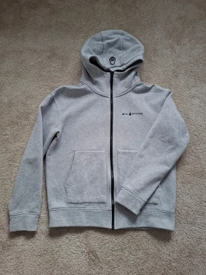 Grå hoodie från Sail Racing - Snygg grå hoodie från Sail Racing med dragkedja framtill och två stora fickor. Märkets logga finns broderad på bröstet och huvan. Tillverkad i mjuk bomullsblandning för skön känsla. Perfekt för en avslappnad och sportig stil.