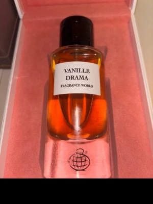 Vanille Drama parfym från Fragrance World - Vanille Drama från Fragrance World är en parfym i en elegant glasflaska med svart kork. Doften har en varm, gyllene färg och flaskan har en modern, stilren design med vit etikett. Perfekt för dig som gillar söta och unika dofter.