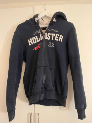 Mörkblå Hollister hoodie med dragkedja - Mörkblå hoodie från Hollister med broderad logga och text på bröstet, dragkedja framtill och två fickor. Mjuk och fluffig insida samt huva med snören. Perfekt för chill dagar och streetwear-stil. Sitter som en M