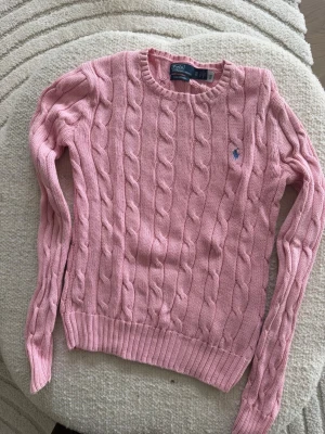 Rosa kabelstickad tröja från Polo Ralph Lauren storlek XS - Snygg rosa kabelstickad tröja från Polo Ralph Lauren med rund halsringning och det klassiska blå broderade logot på bröstet. Tröjan har långa ärmar och ribbade muddar vid ärmslut och nederkant. Perfekt för en trendig och avslappnad look.
