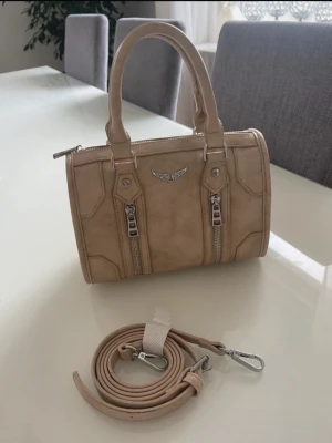 Beige  handväska - Snygg beige handväska zadig voltair dupe från Spanien 💕med två korta handtag och avtagbar axelrem. Väskan har två dekorativa dragkedjor framtill och ett silverfärgat emblem. Tillverkad i konstläder med metalliska detaljer.