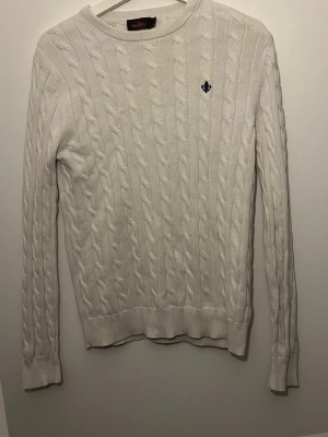 Vit kabelstickad tröja från Morris - Kabelstickad vit tröja från Morris med rund halsringning och diskret broderad logga på bröstet. Tröjan har långa ärmar och ribbade muddar vid ärmslut och nederkant. Perfekt för en klassisk och stilren look.
