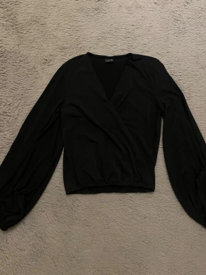 Svart omlottblus från Gina Tricot Stockholmsstil  - Snygg svart omlottblus från Gina Tricot i storlek XS. Blusen har långärmade ballongärmar och en djup v-ringning. Tillverkad i ett mjukt och lätt tyg som ger en skön känsla. Perfekt att matcha med höga jeans eller kjol.