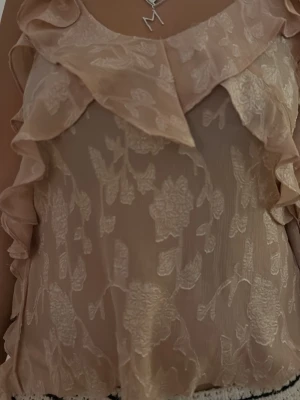 Beige blommig blus med volang - Söt beige blus med blommigt mönster och volangdetaljer. Blusen är ärmlös och har en lätt transparent look med romantiska broderade blommor. Perfekt för dig som gillar en feminin och trendig stil.