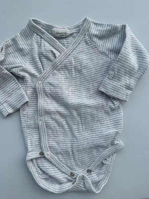 Randig grå omlottbody  - Mysig omlottpyjamas i ljusgrått med tunna mörkgrå ränder. Mjuk och stretchig bomull, tryckknappar framtill och långa ärmar. Perfekt för chill hemma eller som sovplagg.