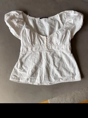 Vit blus från Brandy Melville - Supersöt vit blus från Brandy Melville med korta puffärmar, v-ringning och knappar framtill. Tillverkad i tunn bomull med diskret randigt mönster. Perfekt för en fräsch och somrig look.