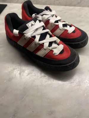 Adidas röda och svarta sneakers - Snygga Adidas sneakers i rött, svart och vitt med klassiska tre ränder på sidan. Skorna har lågt skaft, vita skosnören och en robust svart sula med mönster. Ovandelen är i mocka och textil, perfekt för dig som gillar retrostil. Aldrig använda