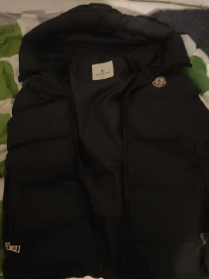 Svart dunväst från Moncler XXL - Snygg svart dunväst från Moncler i storlek M. Västen har en klassisk puffer-design med Moncler-logga på bröstet och dragkedja framtill. Tillverkad i glansigt material med fyllning av dun, perfekt för lager-på-lager-stil. Det står XXL fast egentligen är det M. Falsk storlek 