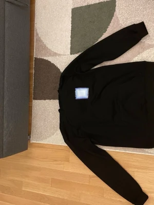 Svart hoodie från Casablanca - Svart hoodie från Casablanca med stor grafisk ruta i blått och vitt på ryggen och texten 'Casa Blanca'. Mjuk bomullskänsla, huva och känguruficka framtill. Perfekt för en chill och stilren look.