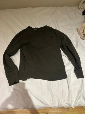 Mörkgrön sweatshirt från Brave Soul - En stilren mörkgrön sweatshirt från Brave Soul med rund halsringning och en liten bröstficka med knapp. Tröjan har breda muddar vid ärmslut och nederkant. Perfekt för en avslappnad look och enkel att matcha med jeans eller joggers. Har även en fin logga på sidan av armen. 