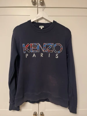 Mörkblå sweatshirt från Kenzo - Säljer en mörkblå vintage sweatshirt från Kenzo med stort broderat Kenzo Paris-tryck i färgglada mönster på bröstet. Tröjan har rund halsringning, ribbade muddar och en avslappnad passform. Perfekt för dig som gillar streetwear och vill sticka ut.