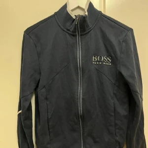 Mörkblå ziptröja från Hugo Boss - Snygg mörkblå ziptröja från Hugo Boss med hög krage och vit logga broderad på bröstet. Tröjan har hel dragkedja framtill och diskreta sömdetaljer. Perfekt för en stilren och sportig look.