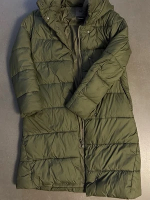 Olivgrön lång dunjacka med huva - Snygg olivgrön dunjacka från Esprit med lång passform och quiltad design. Jackan har dragkedja framtill, två sidofickor och en mysig huva. Perfekt för kalla vinterdagar och riktigt skön att bära.