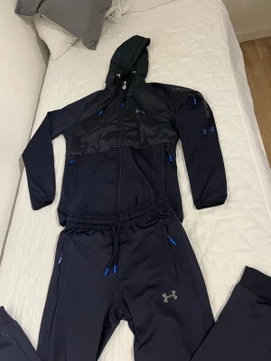 Svart träningsset från Under Armour - Snyggt svart träningsset från Under Armour med matchande hoodie och byxor. Setet har blå detaljer på dragkedjor och snören, samt logga på både jacka och byxa. Materialet är mjukt och stretchigt, perfekt för träning eller chill.