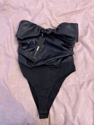 Svart baddräkt med stor rosett - Trendig svart baddräkt från Fashion Nova med en stor, iögonfallande rosett framtill. Axelbandslös modell med glansigt material upptill och klassisk baddräktsform nedtill. Perfekt för dig som vill sticka ut på stranden eller vid poolen.