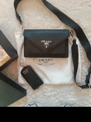 Snyggt väska - Svart axelremsväska  Väldigt elegant. I mycket fint skick. Pyttelite skavd på ett ställe.   Mått 20x13 cm. Djup 5 cm.  Säljes med tillhörande extra liten väska som man kan hänga på väskan.  Box och väskpåse ingår.   Jätte fint material