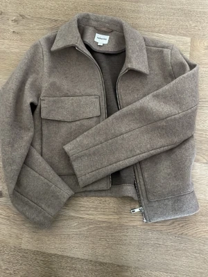 Beige ullovershirt från Kappahl - Kort beige ullkappa från Kappahl med klassisk krage, dragkedja framtill och stora fickor med lock. Kappan har en stilren och minimalistisk design, perfekt för dig som gillar en clean look. Materialet är mjukt och värmande ullblandning. Använd fåtal gånger o kostade 999