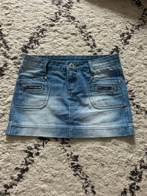 Blå jeanskjol med dragkedjor - Snygg blå jeanskjol i kort modell med två framfickor och coola dragkedjedetaljer. Kjolen har bälteshällor och klassisk knappstängning framtill. Perfekt för dig som gillar en avslappnad och trendig stil.