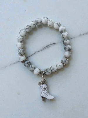 Vitt pärlarmband med bootsberlock - Snyggt armband med vita och gråmelerade pärlor, elastisk tråd och en charmig bootsberlock i vitt med silverdetaljer. Perfekt accessoar för dig som gillar unika detaljer och westerninspirerad stil.