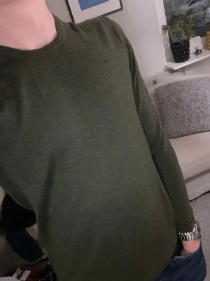 Olivgrön stickad tröja J.Lindeberg - Snygg olivgrön stickad tröja från J.Lindeberg med rund halsringning och diskret logga på bröstet. Tröjan har långa ärmar och ribbade muddar vid ärmslut och nederkant. Perfekt till lager på lager och enkel att matcha.