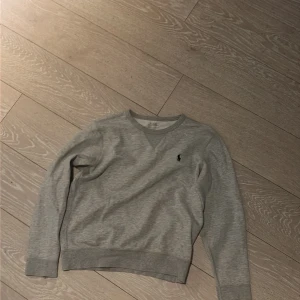 Grå sweatshirt från Polo Ralph Lauren - Klassisk grå sweatshirt från Polo Ralph Lauren med rund halsringning och den ikoniska svarta logotypen broderad på bröstet. Tröjan har ribbade muddar vid ärmslut och nederkant, perfekt för en avslappnad och stilren look.