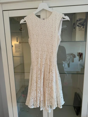 Vit spetsklänning ärmlös - Säljer en söt vit spetsklänning med rund halsringning och ärmlös design. Klänningen har ett vackert blommigt spetsmönster och en kort, utsvängd kjol. Perfekt för sommarens alla tillfällen!