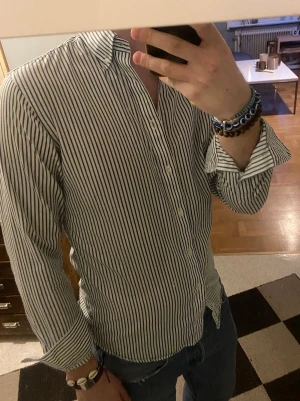 Randig vit skjorta från John Henric  - Snygg vit skjorta med smala gröna ränder och klassisk krage från John Henric. Skjortan har lång ärm och knäppning framtill. Perfekt till jeans eller chinos för en clean look. Storlek 38 passar S